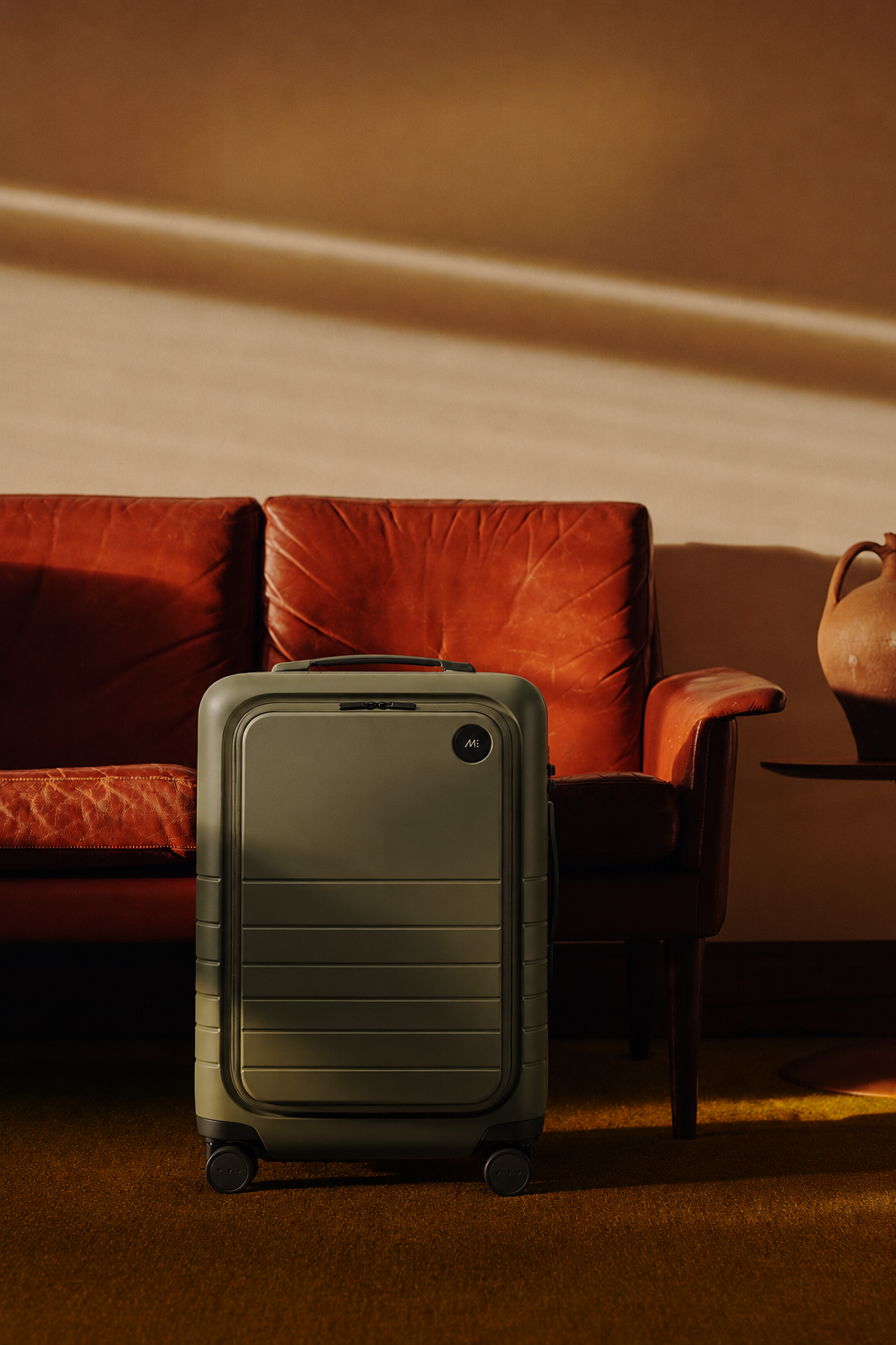 Carry-on campaign visual — olive suitcase by a leather sofa